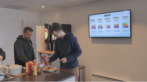 2 men in an office kitchen preparing to eat, and a digital screen behind him
