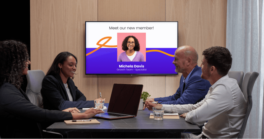 A group of people in a meeting room watching a welcome screen introducing a new team member.